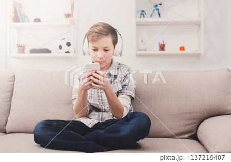 Teenager playing online game on mobile phone and listening to music in earphones at home. Gadget addiction concept Teenager playing online game on mobile phone and listening to music in earphones at home. Gadget addiction concept 137219407