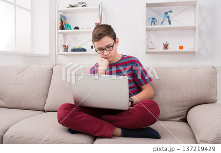 Pensive boy with laptop computer on sofa at home. Child doing homework, looking for information on the internet, typing on keyboard 137219429