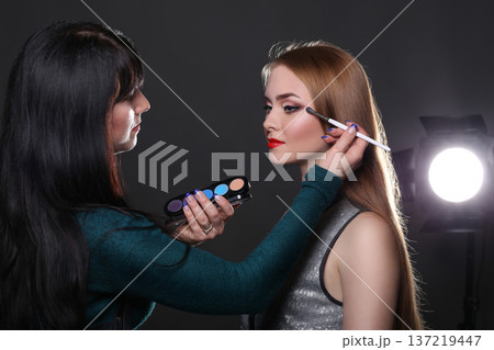 Backstage, fashion shooting. Make-up artist working with beauty model, applying eyeshadow, dark studio background 137219447