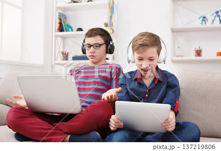 Technology, boy friendship and communication concept - two cute teenagers using tablets, playing games, talking and smiling while sitting on the couch in living room at home Technology, boy friendship and communication concept - two cute teenagers using tablets, playing games, talking and smiling while sitting on the couch in living room at home 137219806