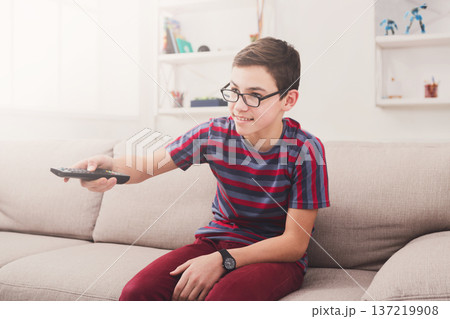 Teenager boy watching television, pointing with remote conroller while sitting on couch in living room at home Teenager boy watching television, pointing with remote conroller while sitting on couch in living room at home 137219908