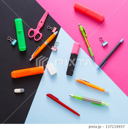 Various stationery supplies are meticulously arranged on a black, pink, and blue surface. Items include pens, markers, scissors, and paper clips, showcasing a vibrant workspace. Various stationery supplies are meticulously arranged on a black, pink, and blue surface. Items include pens, markers, scissors, and paper clips, showcasing a vibrant workspace. 137219937