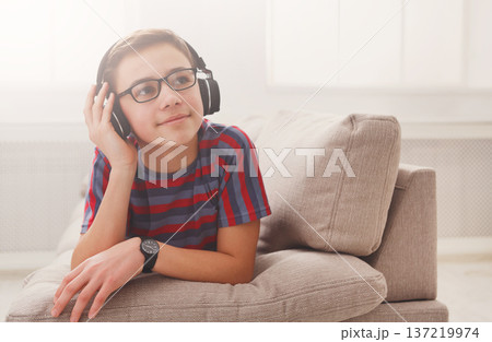 Teenager enjoy music - portrait of dreamy kid in headphones while lying on sofa indoors. Relax time - teenage boy listen to music on couch at home 137219974