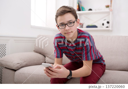 Teenage boy at home with mobile phone, seats on sofa in living room Teenage boy at home with mobile phone, seats on sofa in living room 137220060