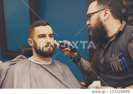 Barber styling beard with trimmer at barbershop. Stylish hairdresser in male hair salon 137220098