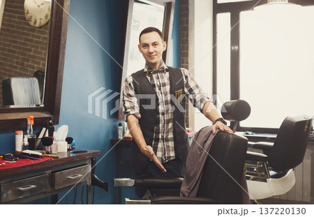 Young elegant barber pointing on a chair, looking at camera and smiling. Male hairstylist inviting customer to have a seat on the chair at barbershop 137220130