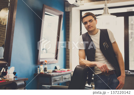 Young barber waits for client, looking at camera. Male hairstylist in barbershop Young barber waits for client, looking at camera. Male hairstylist in barbershop 137220142