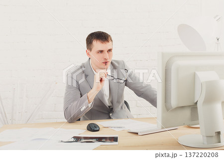Young businessman working in white modern office interior, thoughtful, sitting at computer table. Office worker with eye glasses, job in internet. High key, soft tone. Business man in modern office. 137220208