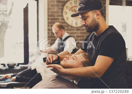 Male barbershop candid image. Styling beard to client. Stylish hairdressers in men hair salon Male barbershop candid image. Styling beard to client. Stylish hairdressers in men hair salon 137220289