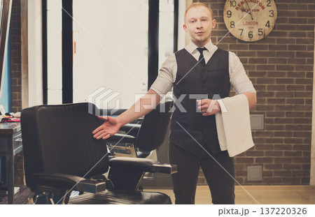 Young elegant barber pointing on a chair, looking at camera and smiling. Male hairstylist inviting customer to have a seat on the chair at barbershop 137220326