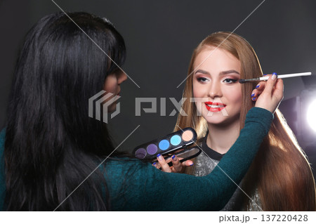 Backstage, fashion shooting. Make-up artist working with beauty model, applying eyeshadow palette, dark studio background 137220428