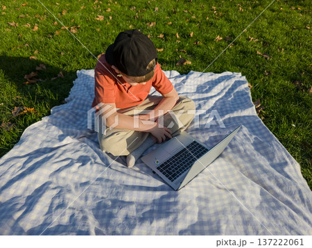 Boy Using Laptop Outdoors in Park, Child Studying Online on Blanket in Nature, Remote Learning Concept Boy Using Laptop Outdoors in Park, Child Studying Online on Blanket in Nature, Remote Learning Concept 137222061