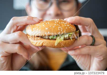 Cropped shot view of young Asian woman holding a hamburger in hands before eating. A hamburger is a sandwich consisting of a cooked meat patty on a bun or roll. 137223479
