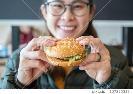 Portrait of young Asian woman holding a hamburger in hand before eating. A hamburger is a sandwich consisting of a cooked meat patty on a bun or roll. Portrait of young Asian woman holding a hamburger in hand before eating. A hamburger is a sandwich consisting of a cooked meat patty on a bun or roll. 137223535