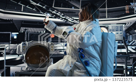 Cheerful astronaut takes selfies with smartphone inside orbital station while traveling toward Mars on exploration mission. Woman takes photos with phone during planet research expedition 137223575