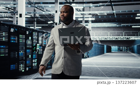 Data center system administrator tasked with reviewing code for AI automation scripts. African american man walking in server hub running diagnostics on equipment rigs, camera A 137223607
