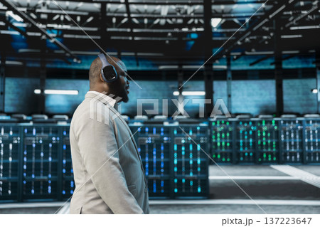 Engineer working in data center, listening music and overseeing infrastructure mainframes. Employee wearing audio headphones walking in server farm delivering web content to online customers Engineer working in data center, listening music and overseeing infrastructure mainframes. Employee wearing audio headphones walking in server farm delivering web content to online customers 137223647