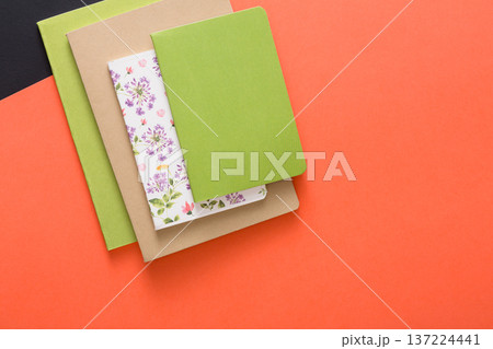 Three notebooks in different colors and styles are neatly stacked on a vibrant orange background. The arrangement showcases a blend of floral patterns and smooth textures, promoting inspiration. 137224441