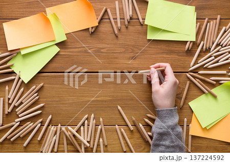 A hand holds a wooden pencil surrounded by scattered colorful notes and pencils on a wooden table. The setup suggests an active brainstorming session, full of ideas and creativity. A hand holds a wooden pencil surrounded by scattered colorful notes and pencils on a wooden table. The setup suggests an active brainstorming session, full of ideas and creativity. 137224592