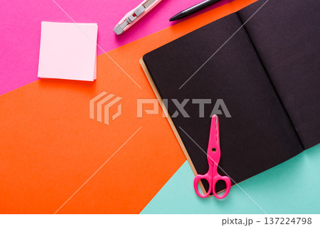 A workspace is organized with a variety of colorful stationery items. Scissors, a notebook, sticky notes, and a pen are neatly arranged on bright paper surfaces, creating a lively atmosphere. A workspace is organized with a variety of colorful stationery items. Scissors, a notebook, sticky notes, and a pen are neatly arranged on bright paper surfaces, creating a lively atmosphere. 137224798