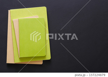 Three notebooks of varying sizes and shades of green are neatly stacked on a dark surface. The notebooks feature textured covers, creating a harmonious arrangement that highlights their colors. 137224879