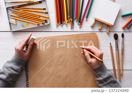 Close-up of artist's workplace - hands with pencil on wooden desktop with blank paper and drawing tools, top view, flat lay Close-up of artist's workplace - hands with pencil on wooden desktop with blank paper and drawing tools, top view, flat lay 137224890