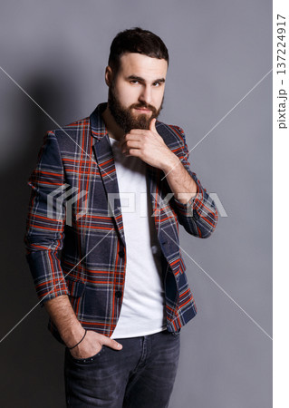 Puzzled young man stroking beard looking at camera. Serious bearded hipster touching his face, gray studio background Puzzled young man stroking beard looking at camera. Serious bearded hipster touching his face, gray studio background 137224917
