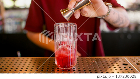 Preparing beverage. Alcohol mixing. Bartender pouring red liquid to glass with ice cubes. Measuring bright cocktail ingredient with jigger. 137230029