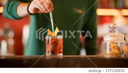 Preparing beverage. Scented cocktail. Barkeeper holding with tweezers decorative figurine orange peel garnishing glass of bright liquid. Preparing beverage. Scented cocktail. Barkeeper holding with tweezers decorative figurine orange peel garnishing glass of bright liquid. 137230035