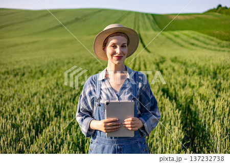 A woman in overalls and a straw hat stands in a green field holding a tablet, representing modern agriculture and technology A woman in overalls and a straw hat stands in a green field holding a tablet, representing modern agriculture and technology 137232738
