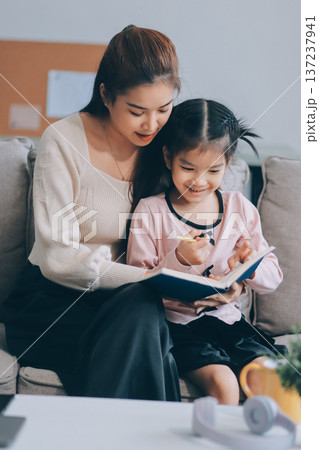 A cheerful Asian mother helps her young daughter do homework and color a book at home, while the father works on a laptop in the background. A cozy, bright, and modern family lifestyle. 137237941