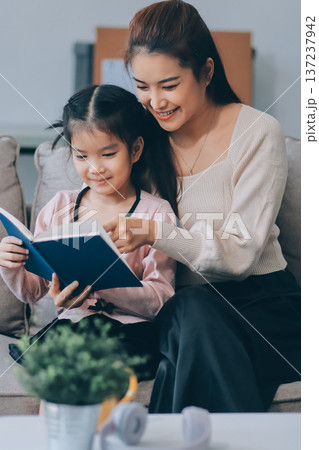 A cheerful Asian mother helps her young daughter do homework and color a book at home, while the father works on a laptop in the background. A cozy, bright, and modern family lifestyle. 137237942