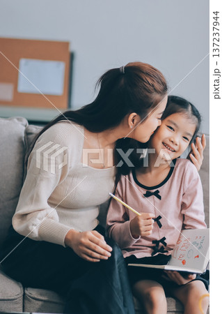 A cheerful Asian mother helps her young daughter do homework and color a book at home, while the father works on a laptop in the background. A cozy, bright, and modern family lifestyle. 137237944