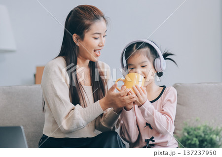 A cheerful Asian mother helps her young daughter do homework and color a book at home, while the father works on a laptop in the background. A cozy, bright, and modern family lifestyle. A cheerful Asian mother helps her young daughter do homework and color a book at home, while the father works on a laptop in the background. A cozy, bright, and modern family lifestyle. 137237950