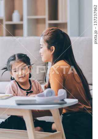 A cheerful Asian mother helps her young daughter do homework and color a book at home, while the father works on a laptop in the background. A cozy, bright, and modern family lifestyle. A cheerful Asian mother helps her young daughter do homework and color a book at home, while the father works on a laptop in the background. A cozy, bright, and modern family lifestyle. 137237962