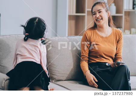 A cheerful Asian mother helps her young daughter do homework and color a book at home, while the father works on a laptop in the background. A cozy, bright, and modern family lifestyle. A cheerful Asian mother helps her young daughter do homework and color a book at home, while the father works on a laptop in the background. A cozy, bright, and modern family lifestyle. 137237980