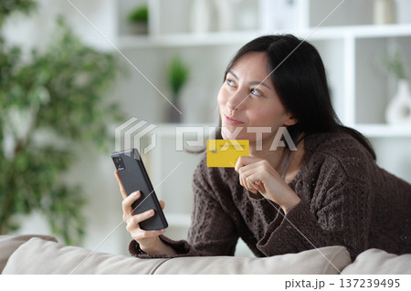 Pensive asian woman holding card and phone at home 137239495