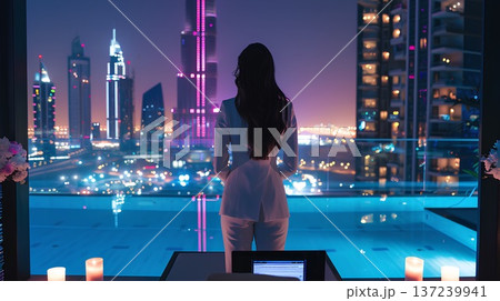 woman in a white suit, standing on the balcony of an ultra-luxury penthouse overlooking dubai city at night with city lights, flowers, and candles around her laptop table.  137239941
