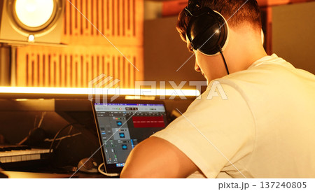 Young Audio Producer Edits Music Track On Laptop, Headphones On. Studio Scene Shows Creative Sound Design, Modern Music Production, Focus On Process. Young Audio Producer Edits Music Track On Laptop, Headphones On. Studio Scene Shows Creative Sound Design, Modern Music Production, Focus On Process. 137240805