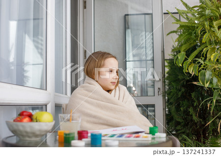 Young child in a warm fleece blanket relaxing on the balcony on cool spring day. Young girl at home. 137241337