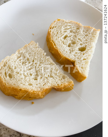 Close-up of two slices of bread placed on a white plate. The scene is minimal and well-lit with a neutral background. Close-up of two slices of bread placed on a white plate. The scene is minimal and well-lit with a neutral background. 137241624