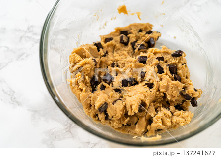 A glass bowl holds chocolate chip cookie dough on a white marble surface. Overhead lighting highlights the thick and textured dough. A glass bowl holds chocolate chip cookie dough on a white marble surface. Overhead lighting highlights the thick and textured dough. 137241627