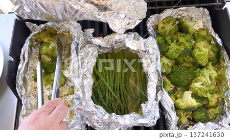 Close-up of a hand holding tongs to flip broccoli and asparagus cooking in foil trays on a gas grill. Natural daylight and rich greens suggest a vibrant summer barbecue scene. 137241630