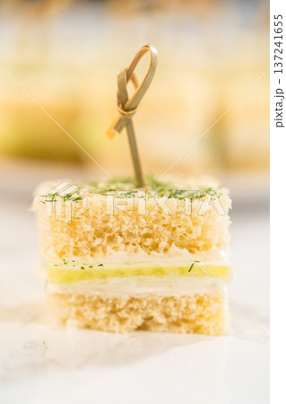 A single mini cucumber sandwich stacked with layers of soft bread and cream cheese, garnished with fresh herbs and served on a marble background, ideal for recipe blogs and tea party features. 137241655