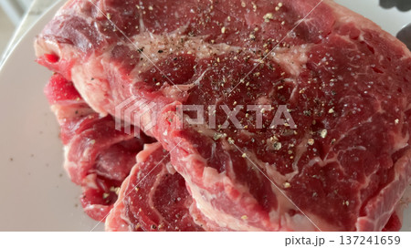 A stack of raw ribeye steaks seasoned with pepper and salt is shown on a white plate. The thick cuts display rich marbling and preparation for grilling. 137241659
