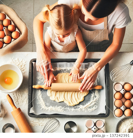 A mother and her young daughter joyfully pipe dough into circles on a baking tray. The little girl's hands assist in shaping the dough, surrounded by flour, eggs, and kitchen utensils in the warm glow 137242391
