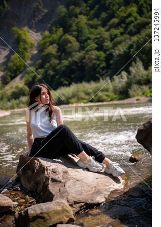 Relaxed Young Woman Resting on Rocks by a Mountain River Relaxed Young Woman Resting on Rocks by a Mountain River 137245994
