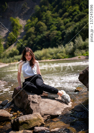 Relaxed Young Woman Resting on Rocks by a Mountain River Relaxed Young Woman Resting on Rocks by a Mountain River 137245996