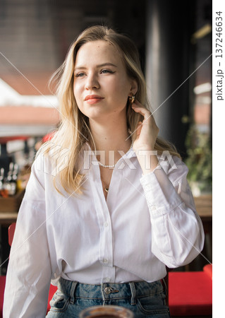 Young woman sitting on a cafe terrace adjusting her hair during a relaxed city break. Urban lifestyle, leisure time, cafe culture, social interaction, everyday elegance and calm modern life. 137246634