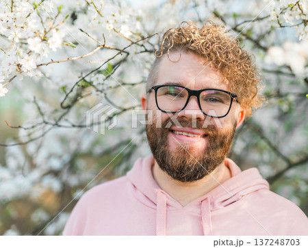 Smiling man portrait among blooming trees during sunny spring day. Joy, confidence, outdoor lifestyle, human connection with nature, peaceful moment during flowering tree blossom season Smiling man portrait among blooming trees during sunny spring day. Joy, confidence, outdoor lifestyle, human connection with nature, peaceful moment during flowering tree blossom season 137248703